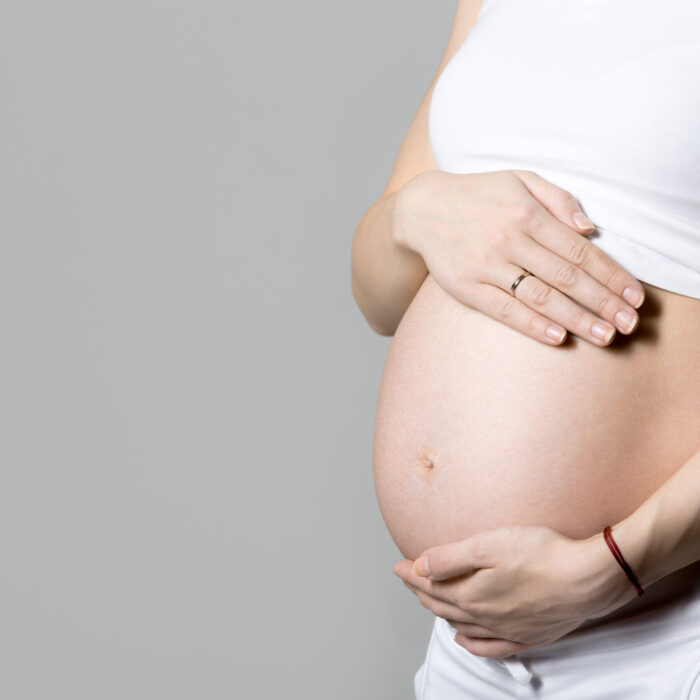 Young pregnant model in tank top touching her belly, gray background, studio, copy space, close-up